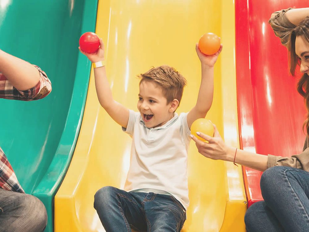 A boy laughing on a slide while his parents toss coloured balls at the playcentre