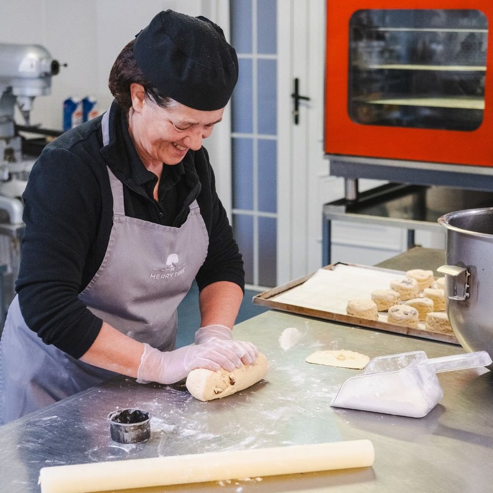 Margaret smiling as she shapes dough on a floured steel bench in the Merry Tree pastry kitchen, with trays of baking behind her